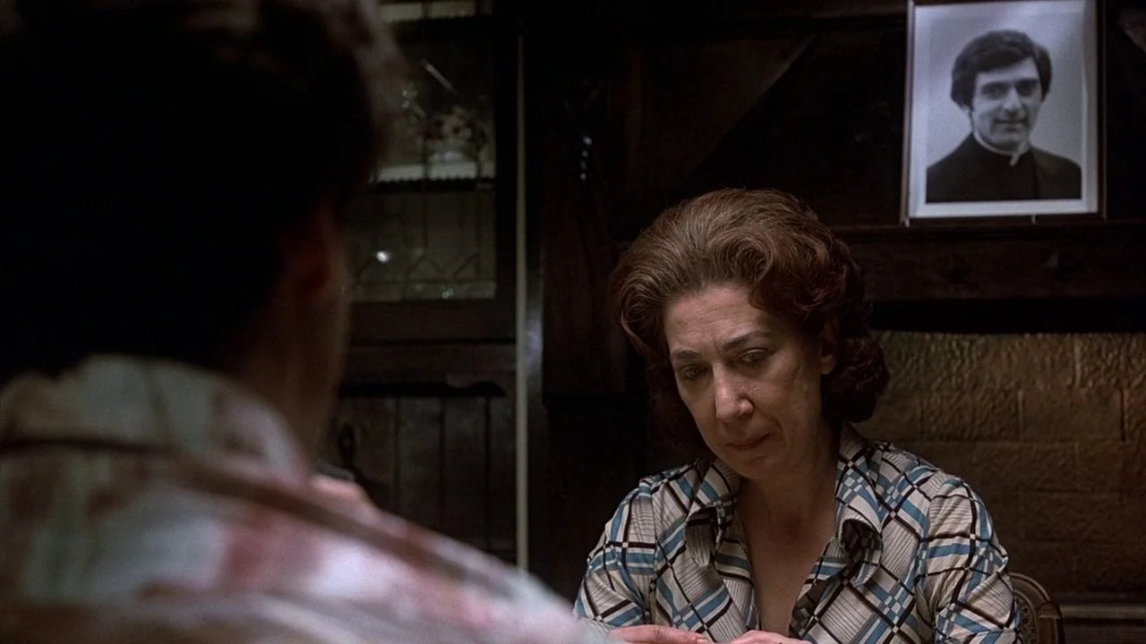 Saturday Night Fever (1977) movie still: medium — A dimly lit room contains a table where an older woman is seated. A framed black and white…