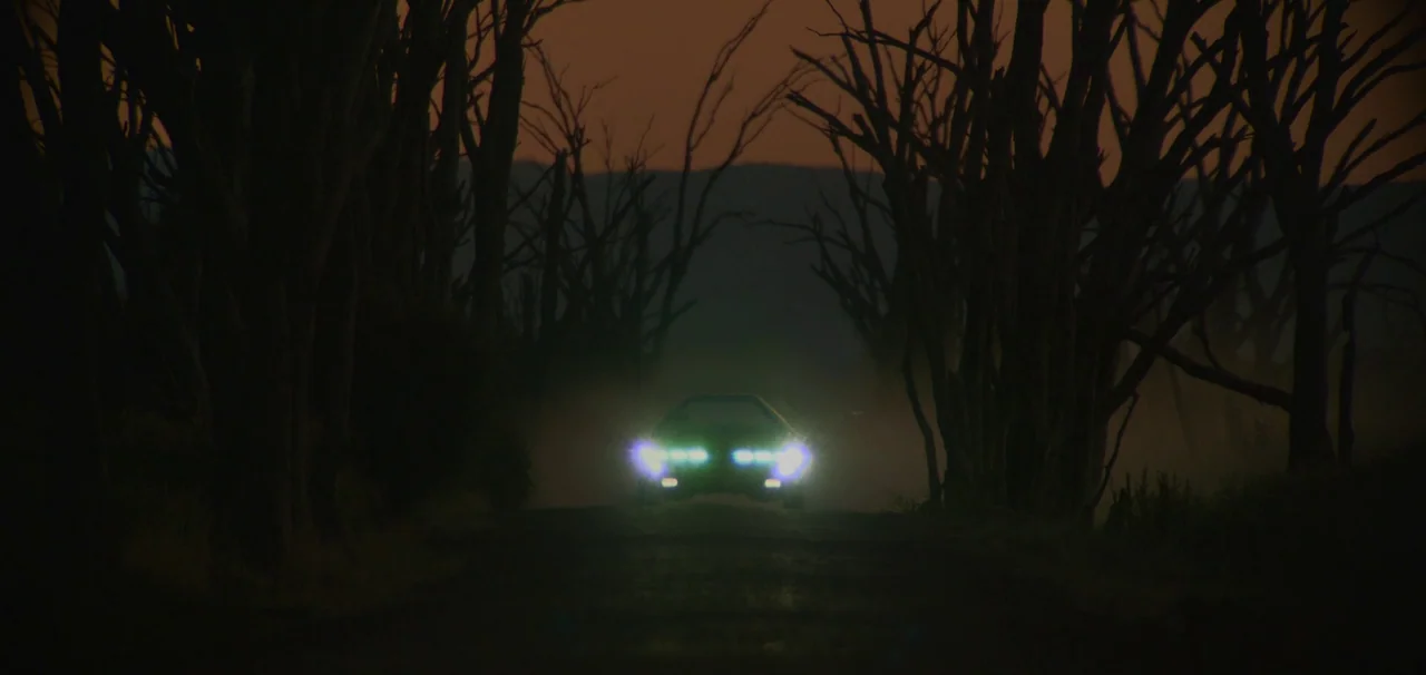 Foe (2023) movie still: wide — A dark, narrow road is flanked by silhouettes of leafless, dead trees against dusk. A car approaches with its bright…