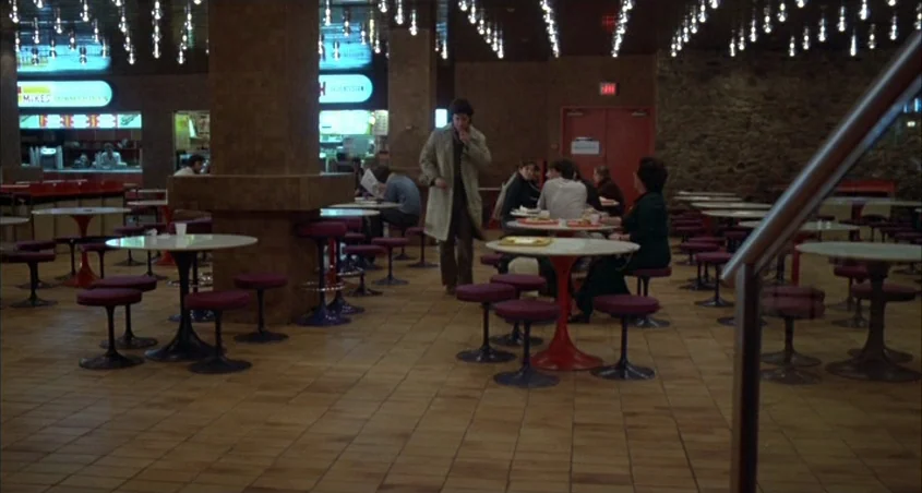 Scanners (1981) movie still: wide — A food court from the 1970s features tiled floors and circular pedestal tables with stools. A man in a trench…