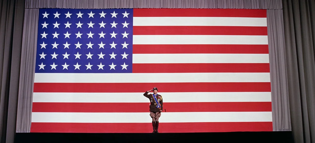Patton (1970) movie still: wide — A man stands centered on a dark stage before an oversized, wall-spanning American flag. He stands perfectly…