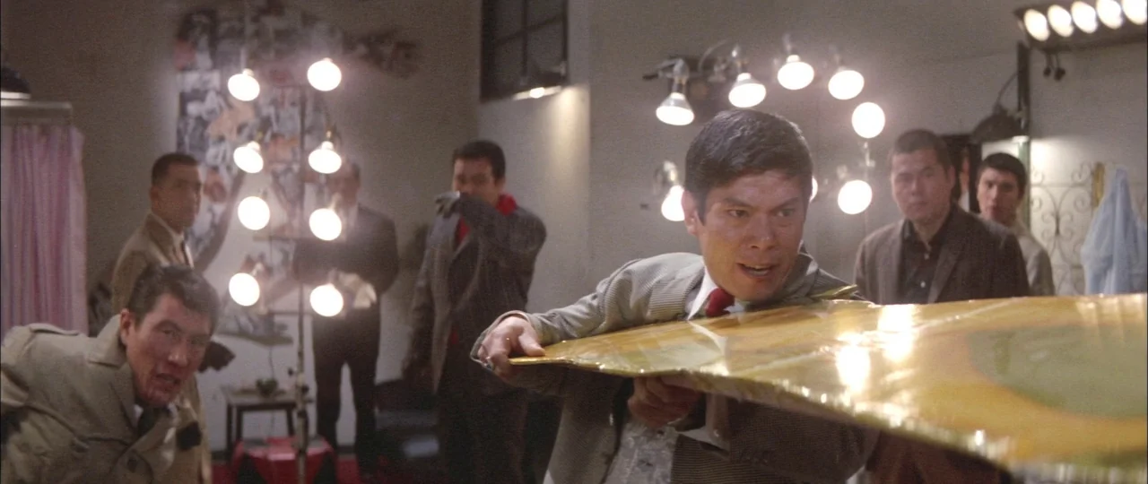 Black Tight Killers (1966) movie still: medium — A crowded dressing room contains multiple people standing around under bright industrial stage…