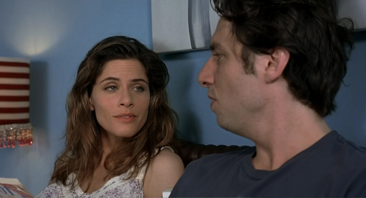Mi Perfecto Ex (2024) movie still: medium — A woman and a man sit in a bedroom with blue walls. The woman faces the man while he is turned in…