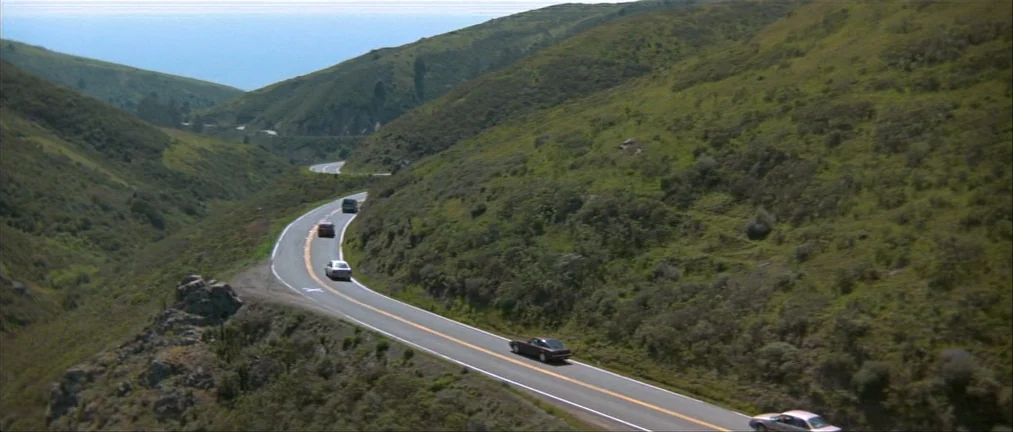 Basic Instinct (1992) movie still: establishing — A winding asphalt road curves through rolling green hills overlooking the ocean. Several vehicles…