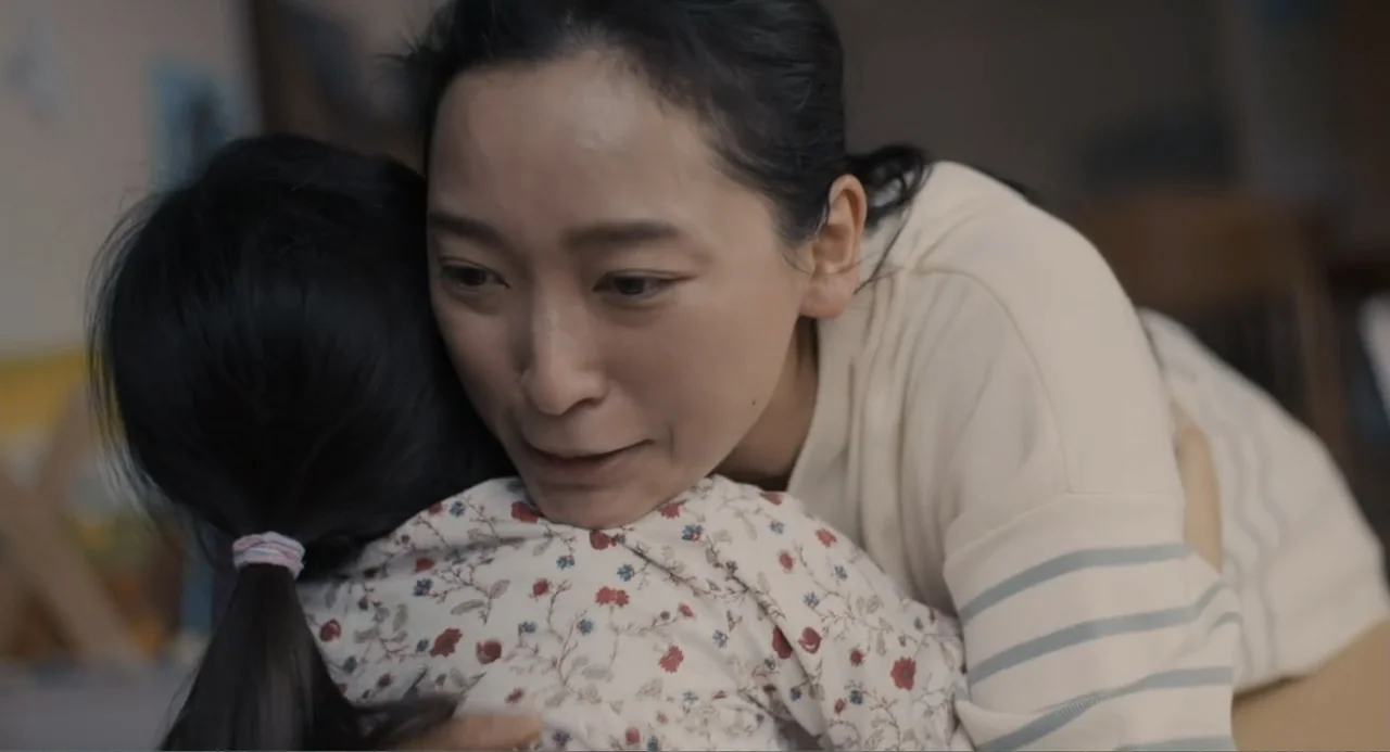 Elles (2018) movie still: closeup — An indoor domestic space with soft lighting and neutral walls. A woman hugs a young child wearing a floral…