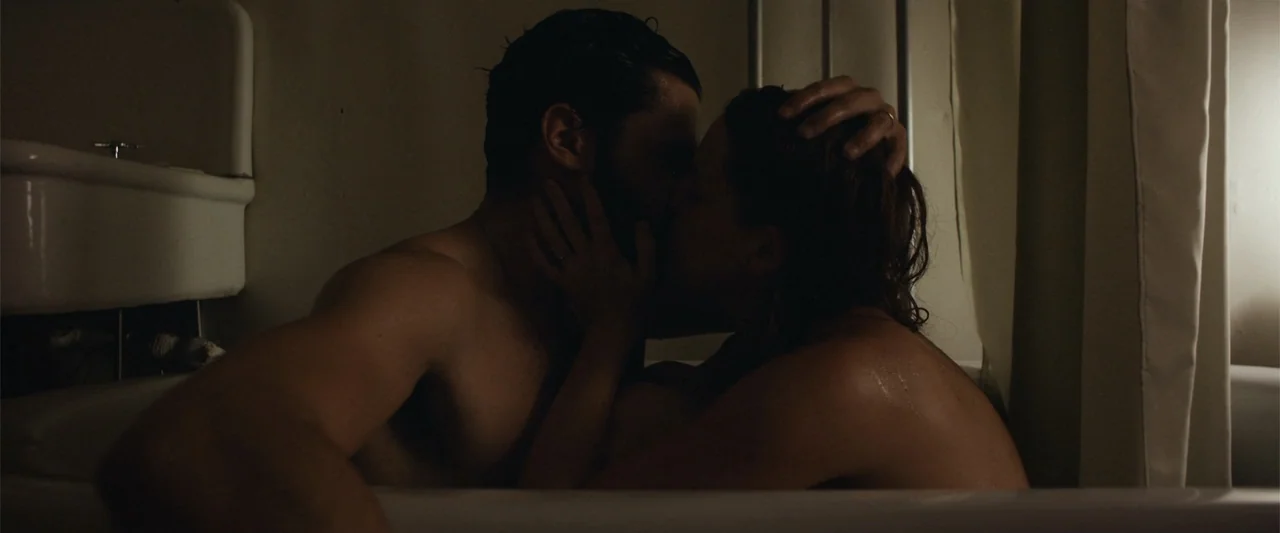 It Comes at Night (2017) movie still: medium — A dimly lit room contains a white porcelain bathtub with two people inside. The couple sits together…