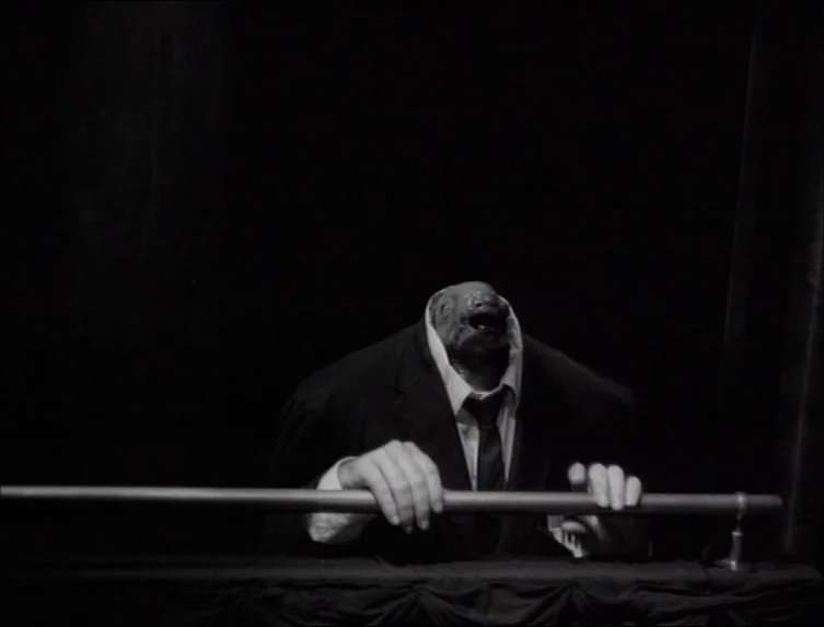Eraserhead (1977) movie still: medium — A dark, stage-like setting features heavy curtains against a deep black background. A creature in a suit…