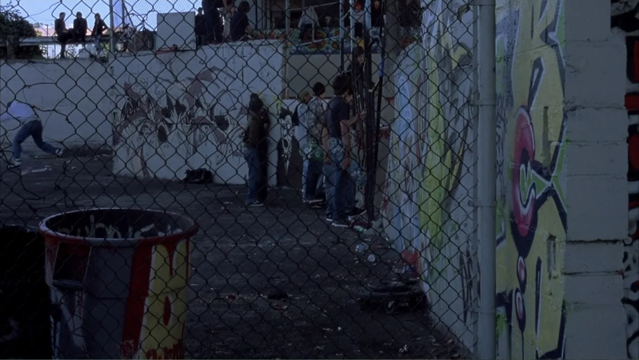 Paranoid Park (2007) movie still: medium — An outdoor area features a chain-link fence foreground and a concrete wall covered in graffiti. A small…