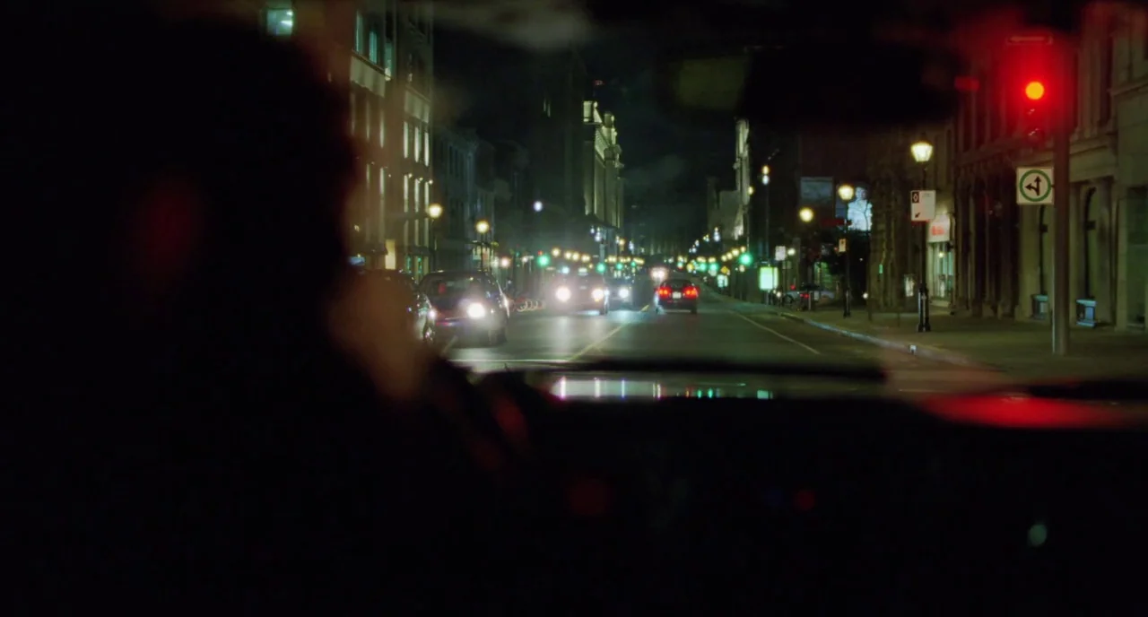 Tom at the Farm (2013) movie still: wide — A nighttime city street view from inside a car stopped at a red light. The road is flanked by stone…