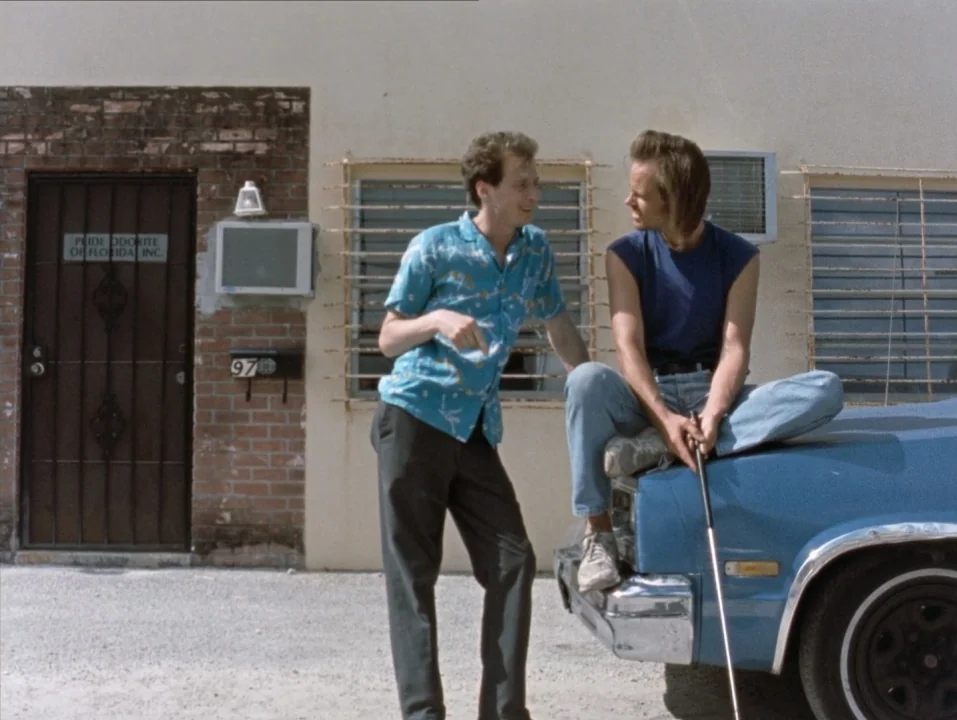 River of Grass (1994) movie still: fullbody — A light-colored stucco building with a brick doorway and barred window sits under daylight. Two men…