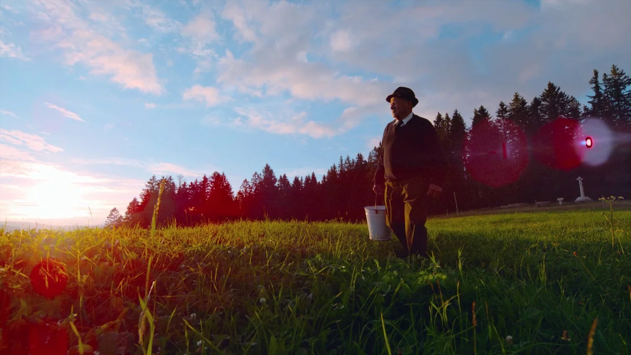 Awaken (2018) movie still: fullbody — A man in a fedora walks through a grassy field towards a line of trees. The sun sets low on the horizon,…