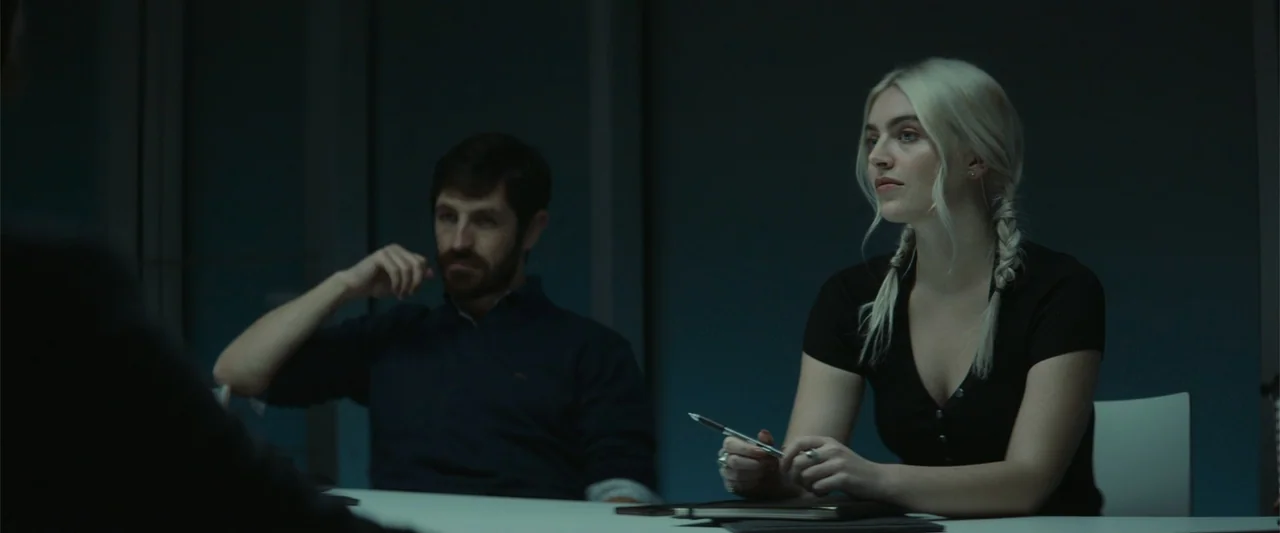 The Cellar (2022) movie still: medium — A modern meeting room features dark wall paneling and a white table surface. A woman and man sit…