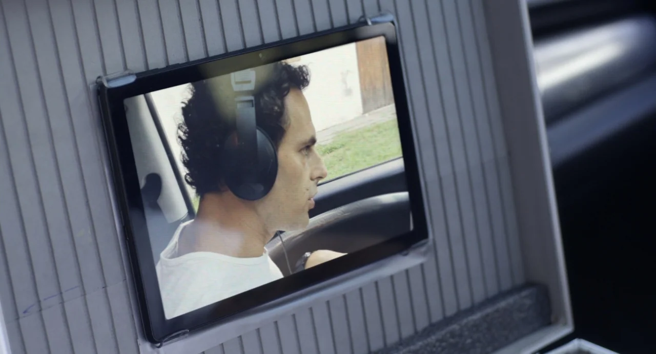 Little White Lies (2010) movie still: medium — A man is captured on a tablet screen displayed inside a gray plastic carrying case. The man sits in…