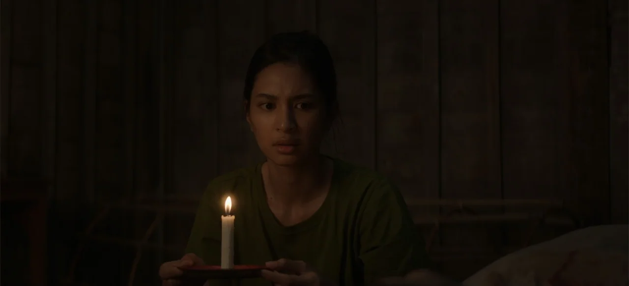 A Thousand Days (2023) movie still: medium — A dimly lit room with walls made of vertical dark wooden planks. A woman sits in the dark holding a lit…