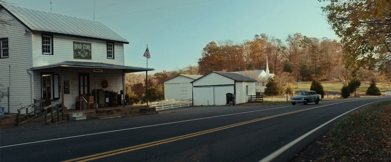 Coming Through the Rye (2016) movie still: wide — A roadside store sits beside an asphalt highway in a rural setting during autumn. A vintage car…
