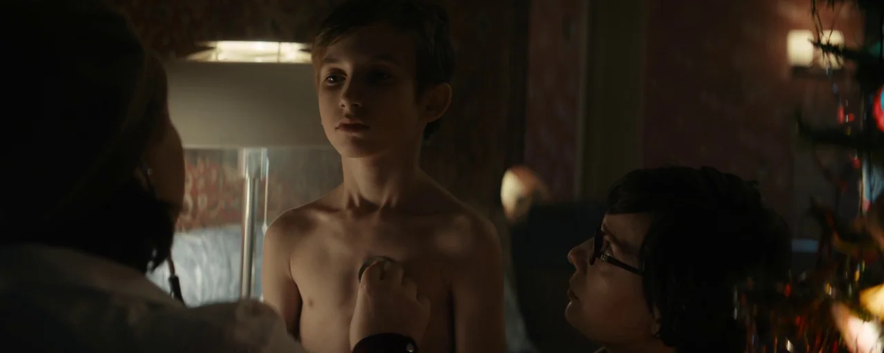 Petrov’s Flu (2021) movie still: medium — A dimly lit room with patterned wallpaper contains a child being examined by a physician. The woman…