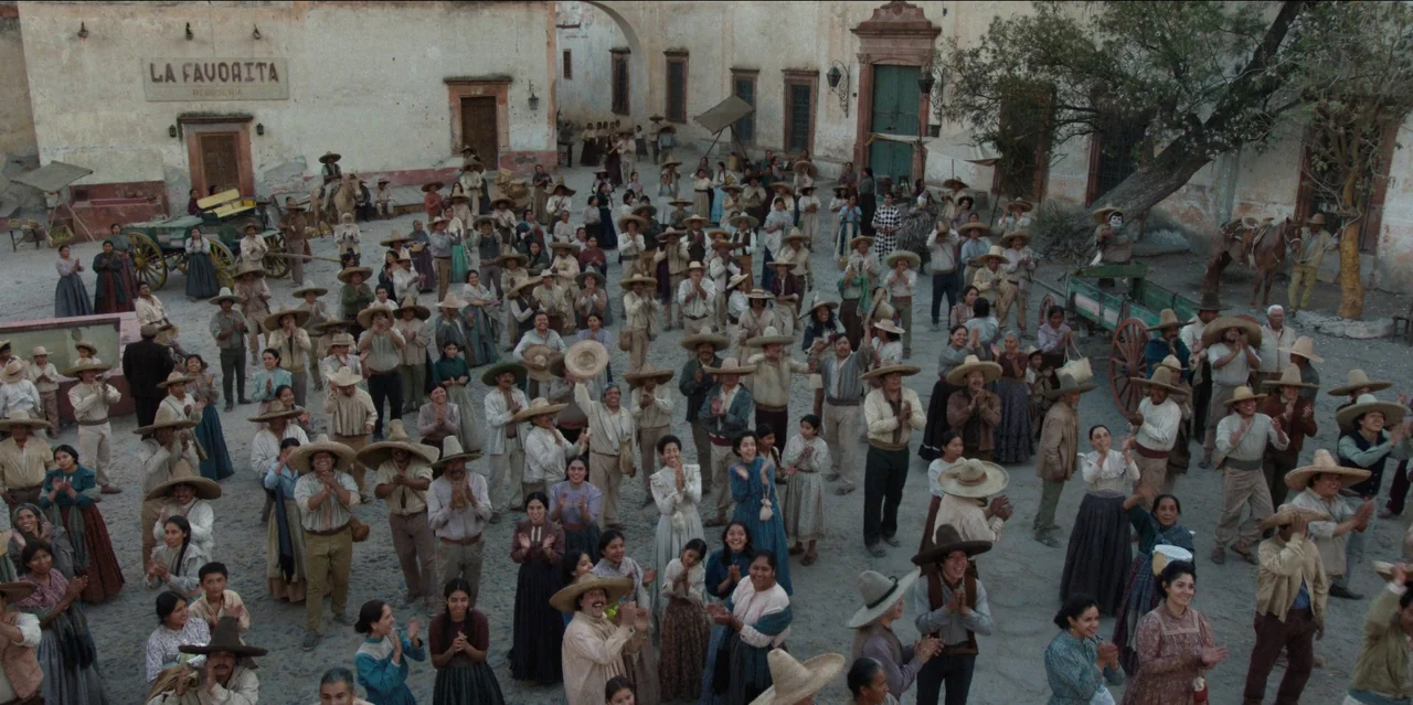 Pedro Páramo (2024) movie still: wide — A dusty town square features beige colonial buildings and a large, gnarled tree. A crowd of people in period…