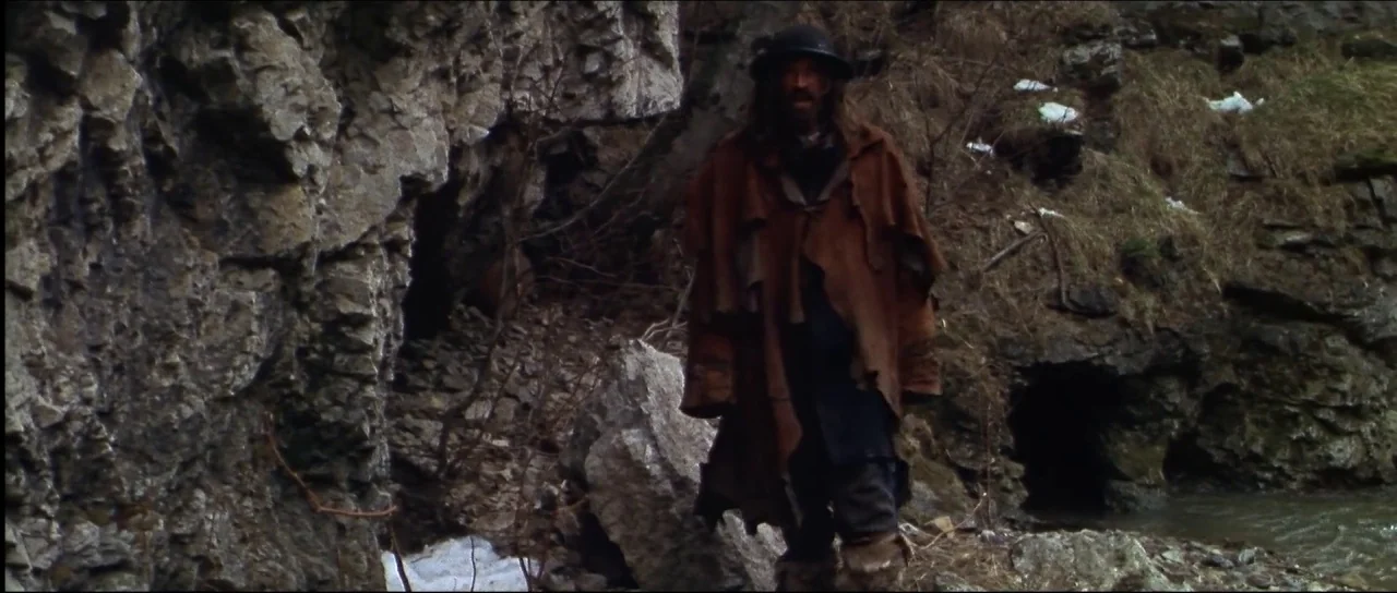 Ravenous (1999) movie still: fullbody — A man stands in a dark rocky ravine with patches of snow on the ground. He wears a heavy leather coat and a…