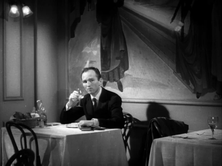 The Seventh Victim (1943) movie still: medium — A restaurant interior features white tablecloths and a large mural on the back wall. A man sits…