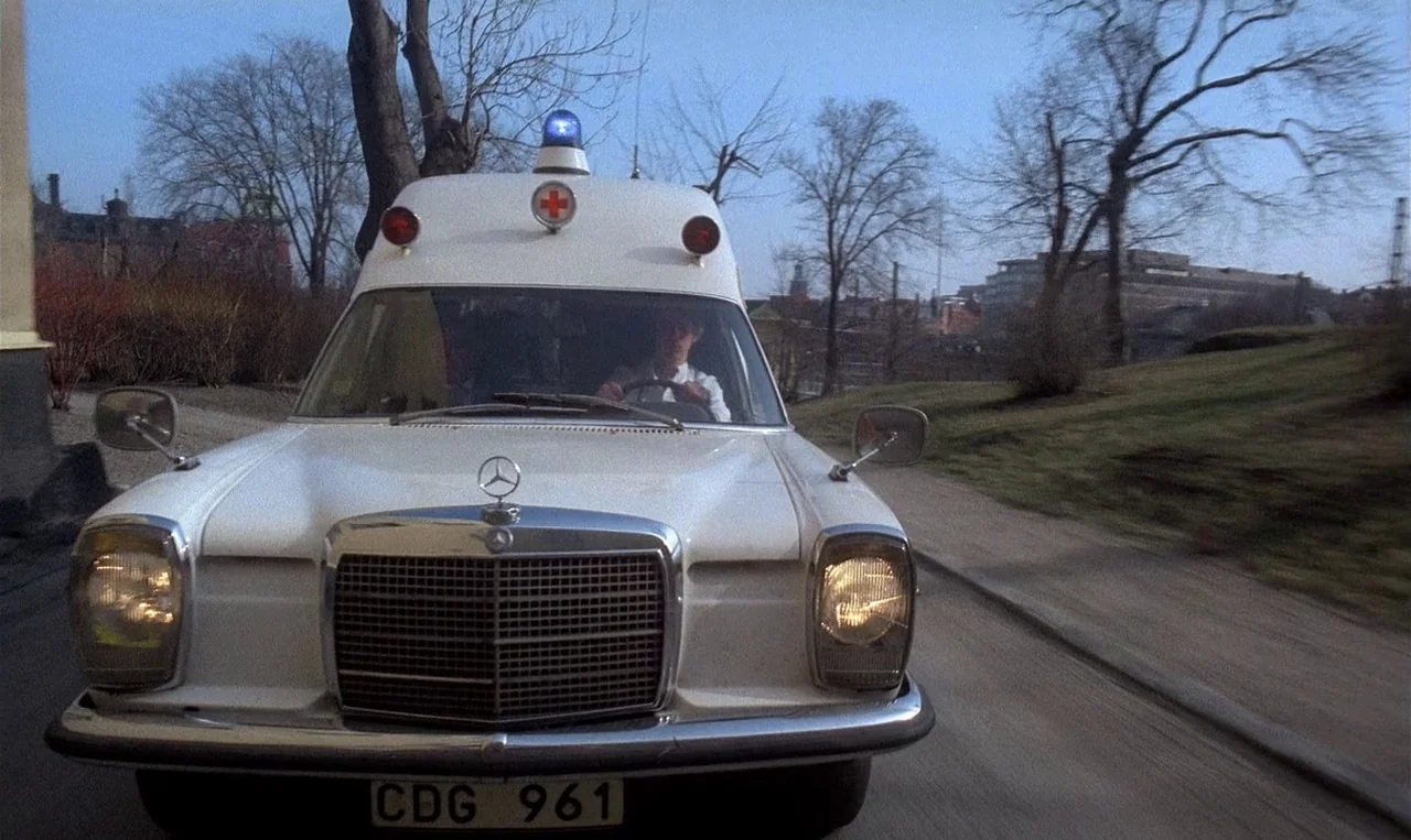 Man on the Roof (1976) movie still: medium — A white vintage ambulance drives along a suburban street with bare trees. The driver keeps his hands on…