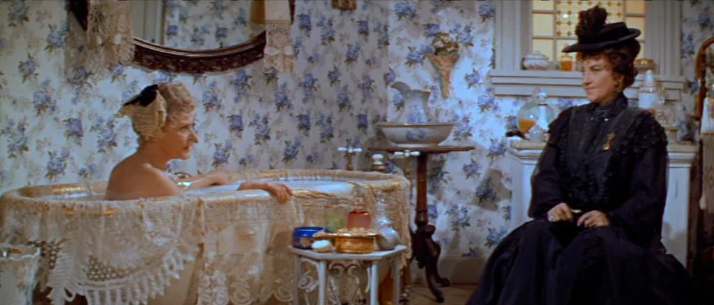 Gigi (1958) movie still: medium — A Victorian-era bathroom features floral blue wallpaper and a lace-covered bathtub. A woman sits in the tub while…
