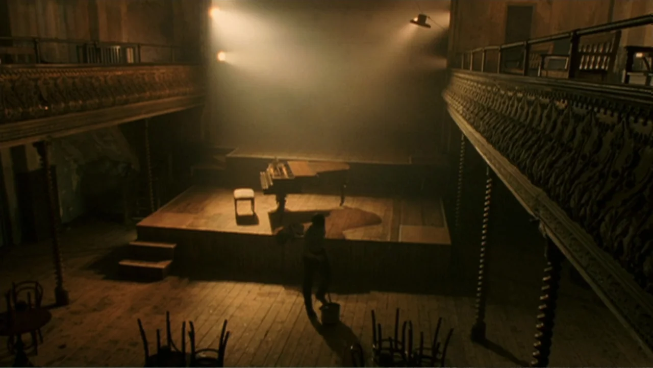 Penelope (2007) movie still: wide — A dark, wood-paneled music hall features a raised stage with a grand piano. A single figure mopping the floor…