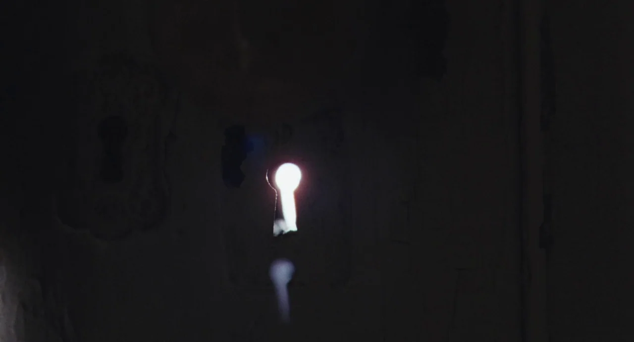 The Giant (2019) movie still: closeup — A dark wooden surface contains a metal keyhole plate. Bright white light spills through the narrow opening…