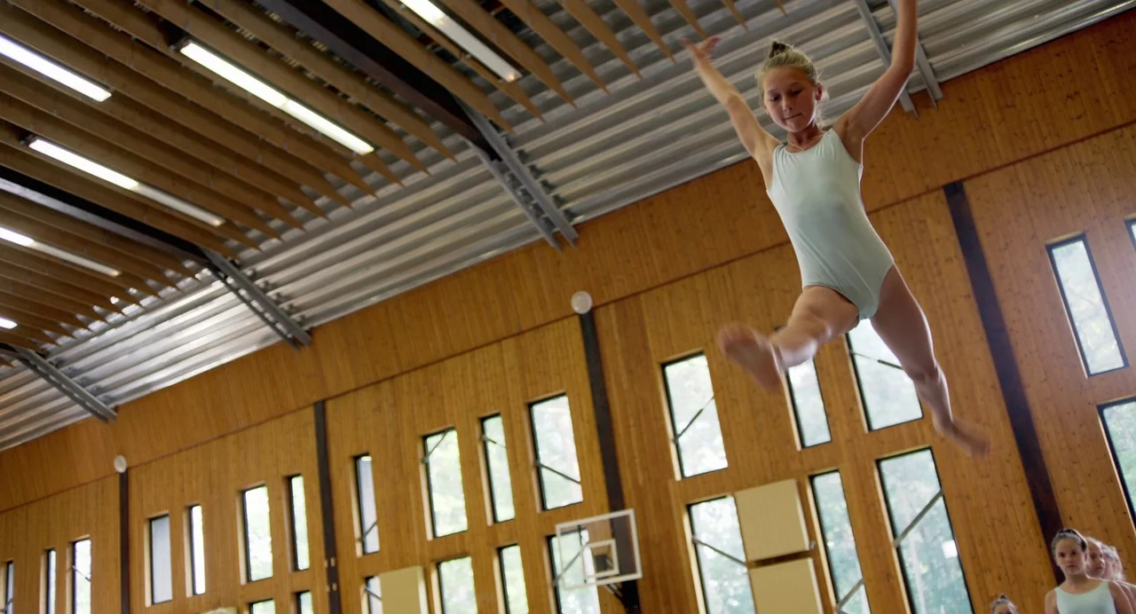 Hatching (2022) movie still: medium — A high-ceilinged gymnasium features warm wood-paneled walls and a series of tall windows. A young female…