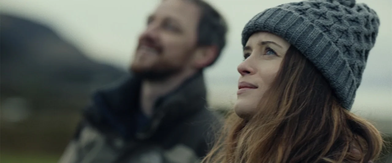 My Son (2021) movie still: medium — An outdoor landscape during a overcast day with rolling hills in the background. A woman in a knit hat and a man…