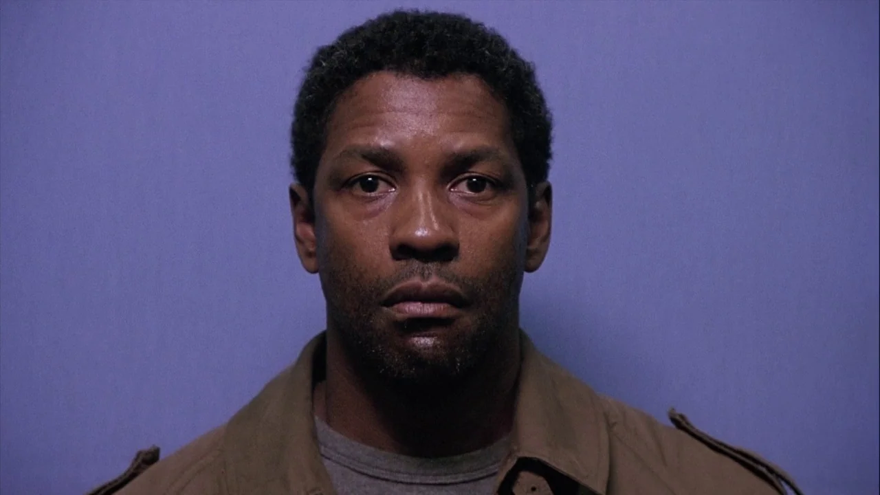 The Manchurian Candidate (2004) movie still: closeup — A man stands against a flat, solid-colored wall during a police mugshot. He looks directly at…