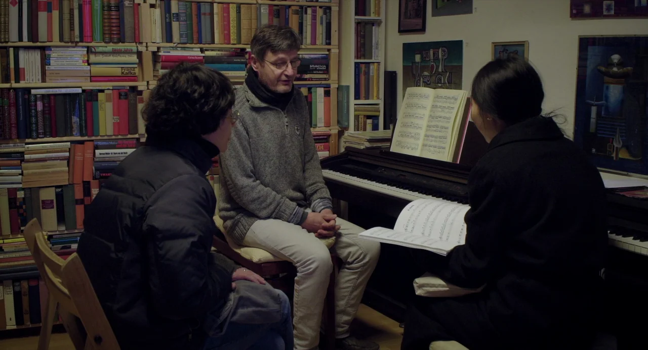 On The Beach At Night Alone (2017) movie still: medium — A room filled with bookshelves holds a piano and three people seated together. The group…