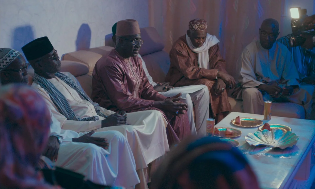 Atlantics (2019) movie still: medium — A group of men in traditional clothing sit on a sofa inside a room. They gather around a small table topped…