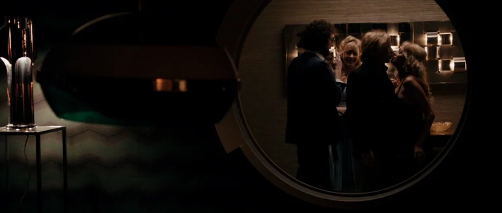 The Look of Love (2013) movie still: medium — A dark, dimly lit 1970s lounge is framed through a circular architectural feature. Three people…