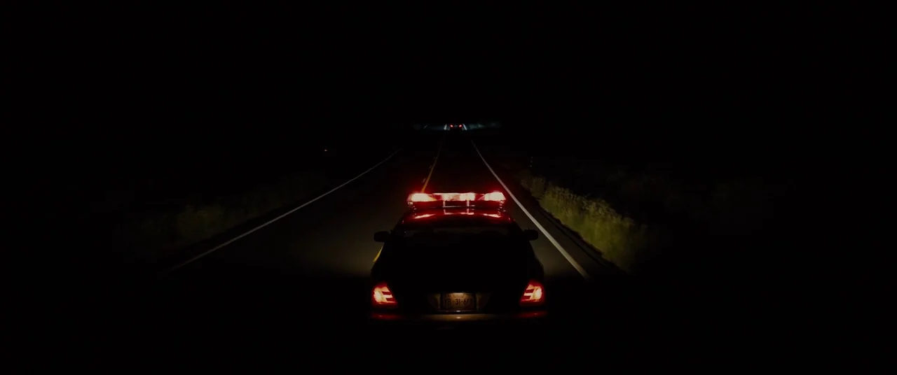 Sicario (2015) movie still: wide — A police car drives down an empty asphalt highway surrounded by absolute midnight darkness. Only the bright red…