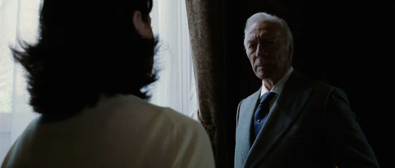 Inside Man (2006) movie still: medium — A dark indoor space features a man standing near a window with sheer curtains. An unidentified woman is…