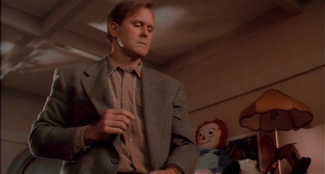 Raising Cain movie still: medium — A dimly lit room contains a soft lamp and a cloth doll in the background. A man in a grey blazer stands in the…