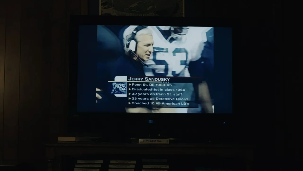 Paterno (2018) movie still: medium — A dark, dimly lit room contains a large television displaying sports statistics. The screen shows a coach…