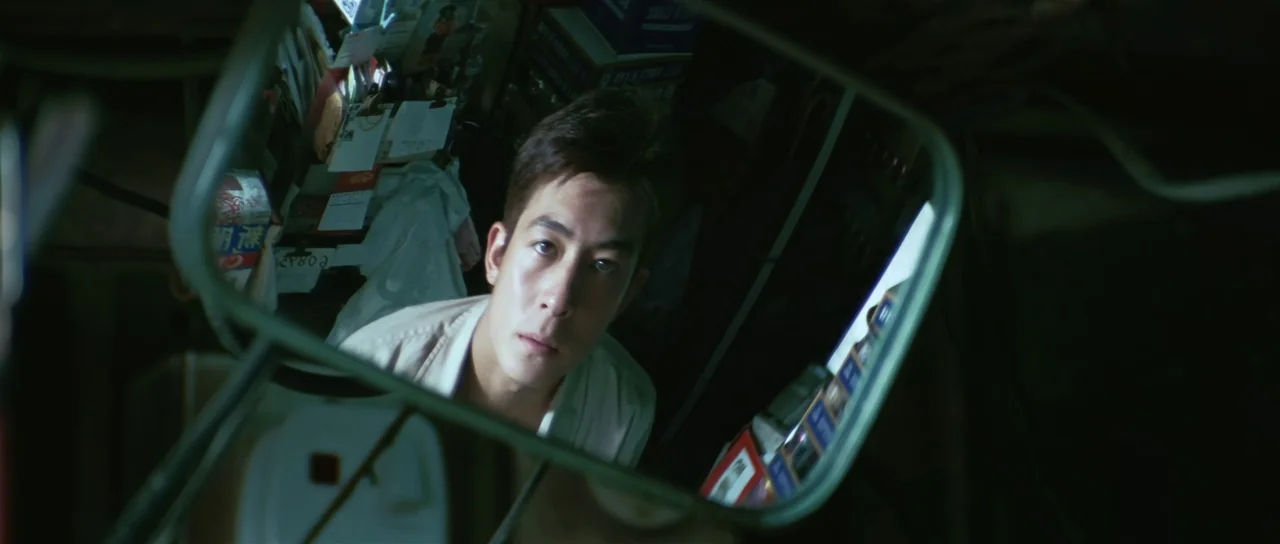 Infernal Affairs (2002) movie still: closeup — A man's face is reflected in a car's rearview mirror. The background behind him is dark and cluttered…