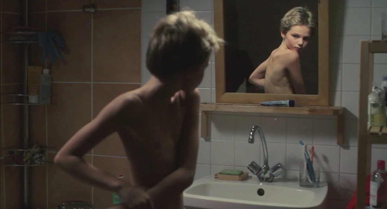 Tomboy (2011) movie still: medium — A tiled bathroom features a sink, mirror, and shelves stocked with toiletries. A young man stands before the…
