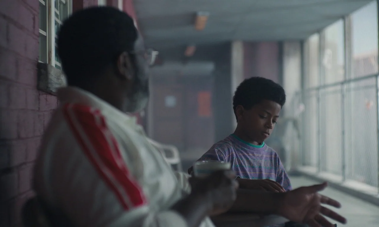 We Grown Now (2023) movie still: medium — A covered concrete walkway with red painted brick walls and chain-link fence sides. An adult man and a…