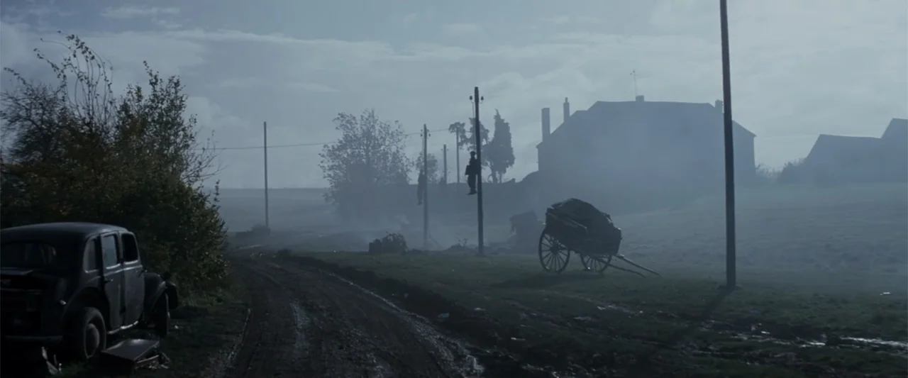 Fury (2014) movie still: wide — A misty, rural dirt road stretches past a farmhouse and wooden utility poles. Two figures hang suspended from the…