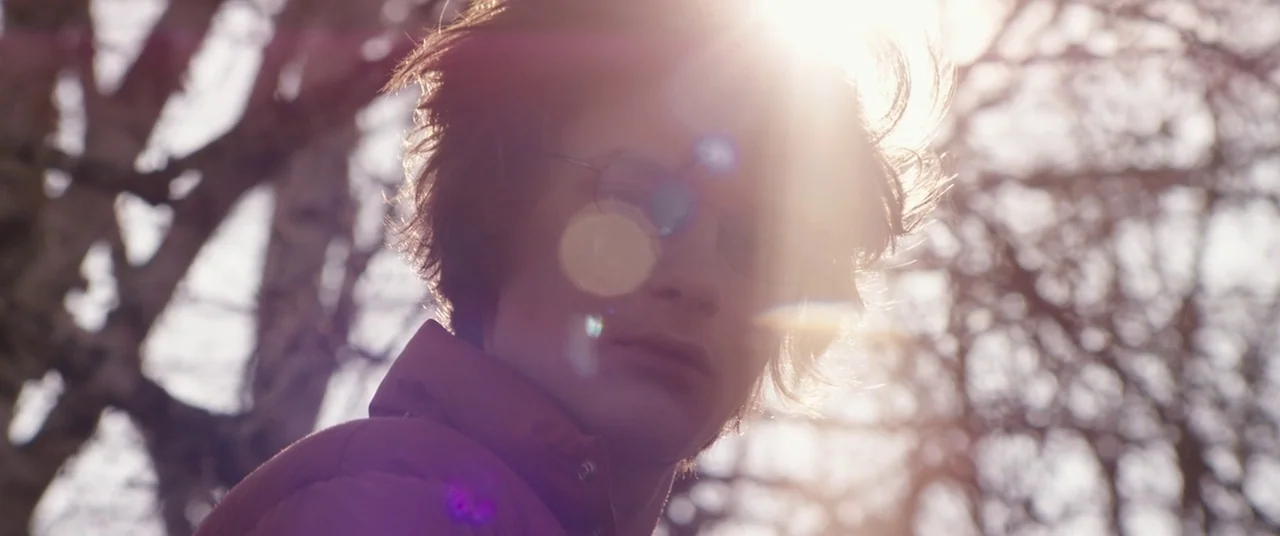Super Dark Times (2017) movie still: closeup — A person stands outdoors in a grove of trees with bare branches during daylight. Sunlight hits the…