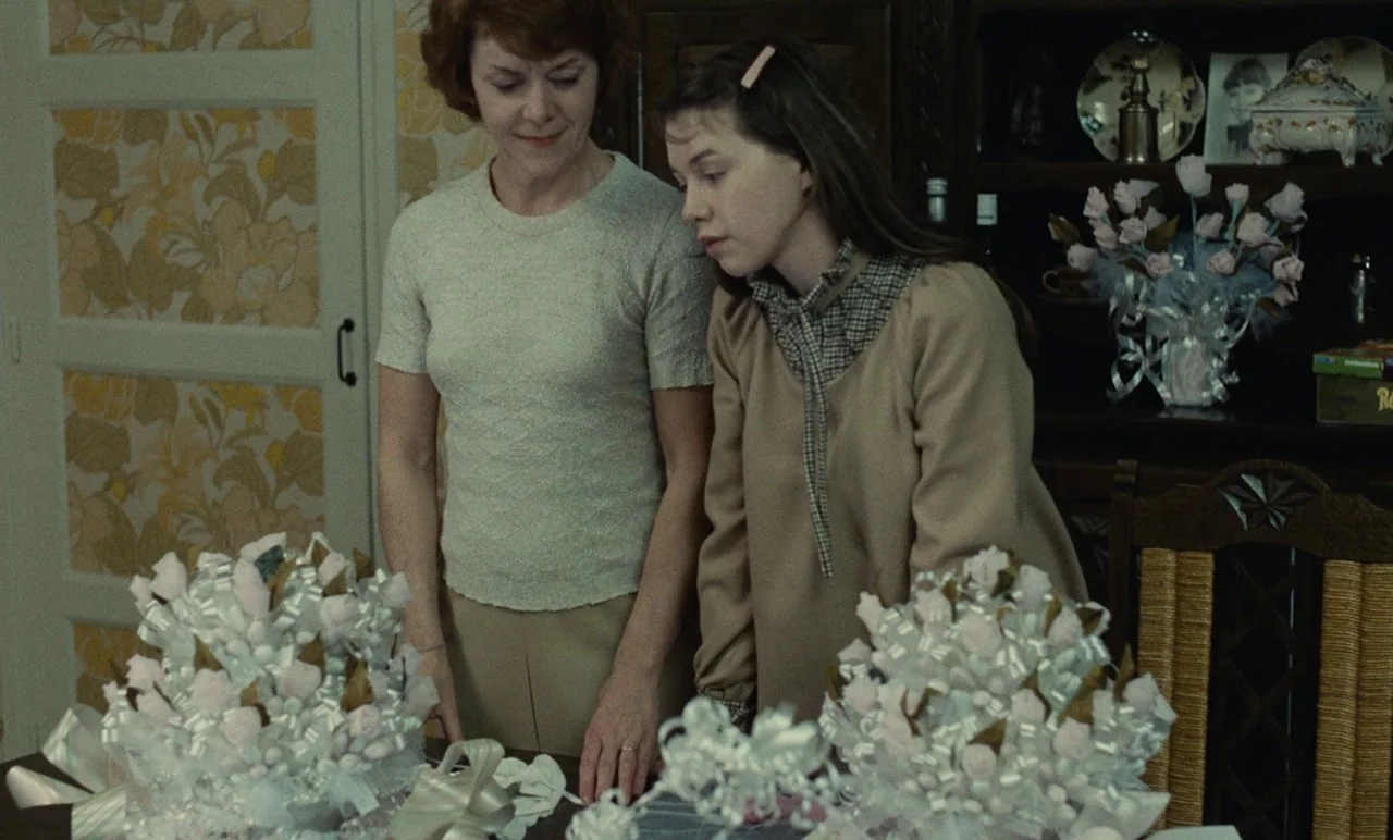 Graduate First (1978) movie still: medium — A residential room features yellow patterned wallpaper and a wooden cabinet with decorative porcelain. A…
