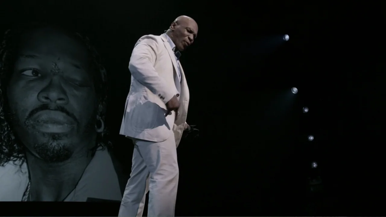 Mike Tyson: Undisputed Truth (2013) movie still: fullbody — A dark, empty stage features a bright spotlight on a man in a white suit. A large…