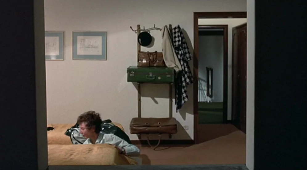 Don’t Look Now (1973) movie still: wide — A hotel room features a wall-mounted coat rack with luggage and hanging coats. A person sits on one of the…