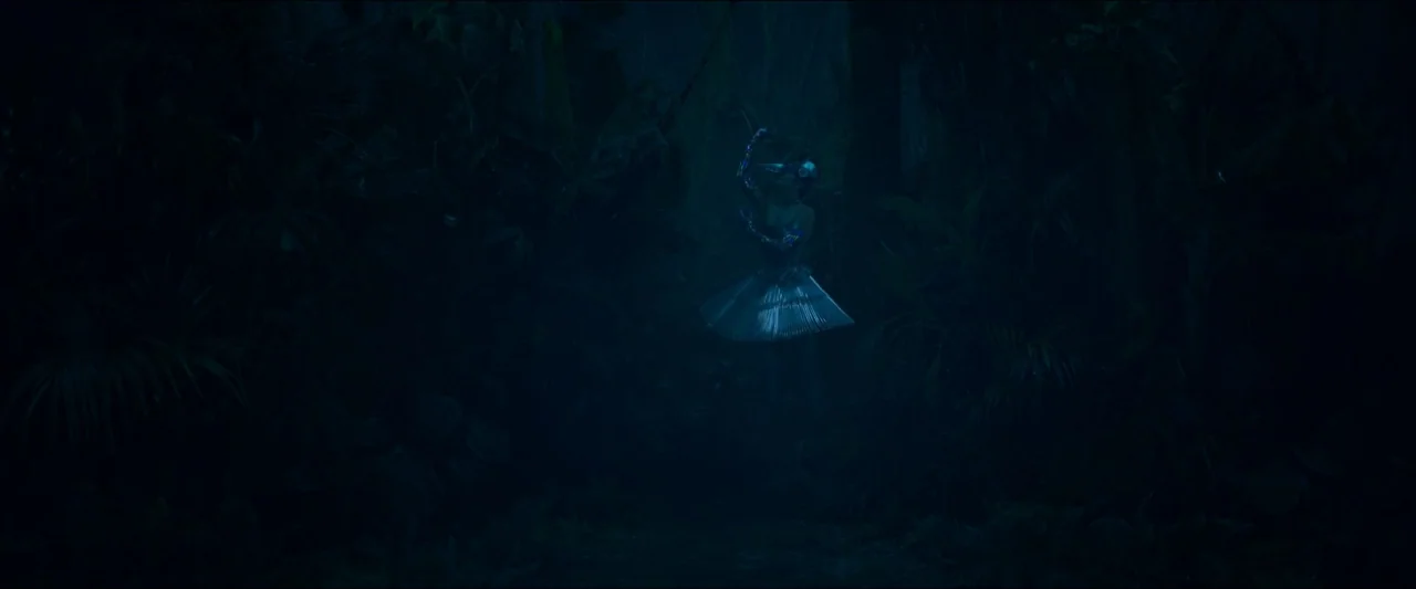 Birds of Paradise (2021) movie still: wide — A dark, dense jungle environment surrounds a single figure in the center frame. The figure wears a…