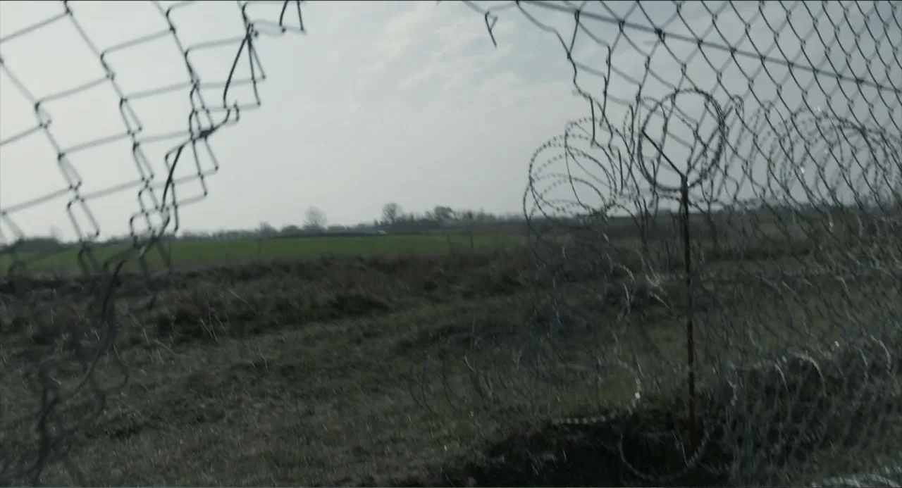 Human Flow (2017) movie still: establishing — An open green field is enclosed by a damaged chain-link fence topped with coils. A large gap in the…