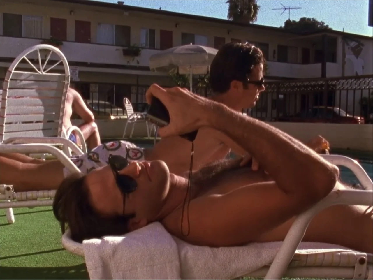 The Living End (1992) movie still: medium — A motel courtyard features a swimming pool with lounge chairs on a green turf deck. Two shirtless men in…