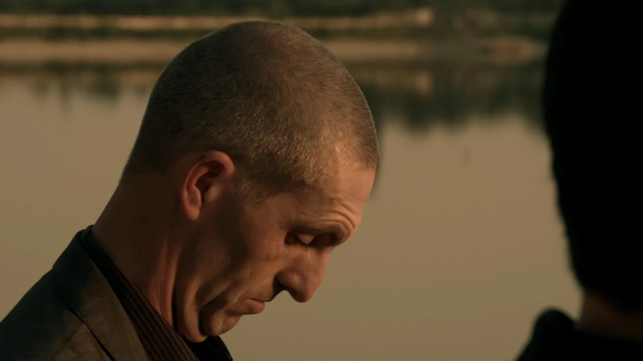 6 Bullets (2012) movie still: closeup — A calm, still riverbank landscape stretches into the soft-focus golden hour background. A man with a buzzcut…