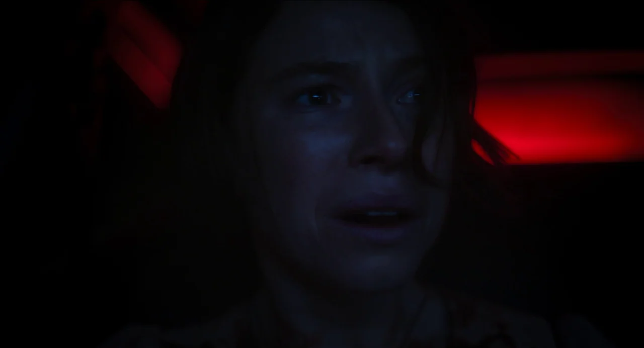 Men (2022) movie still: closeup — An interior of a dark vehicle at night with red neon illumination in back. A woman stares into the distance with…
