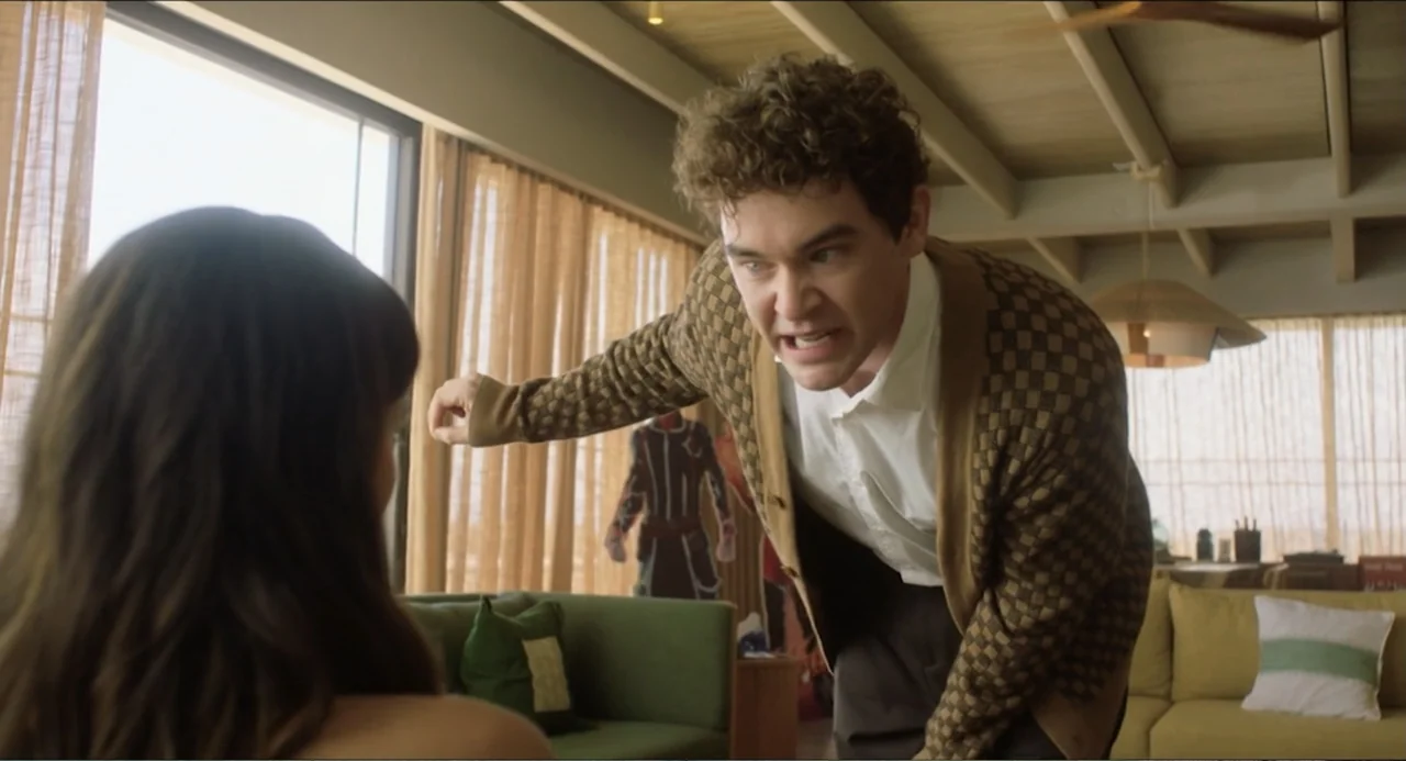 At Midnight (2023) movie still: medium — A living room features a green sofa and large windows with tan curtains. A man in a checkered cardigan…